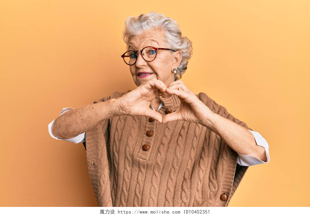 橙色背景前的一个奶奶高个子灰发女人，身穿休闲装，戴着眼镜，带着爱心微笑，手握心形符号。浪漫的概念. 
